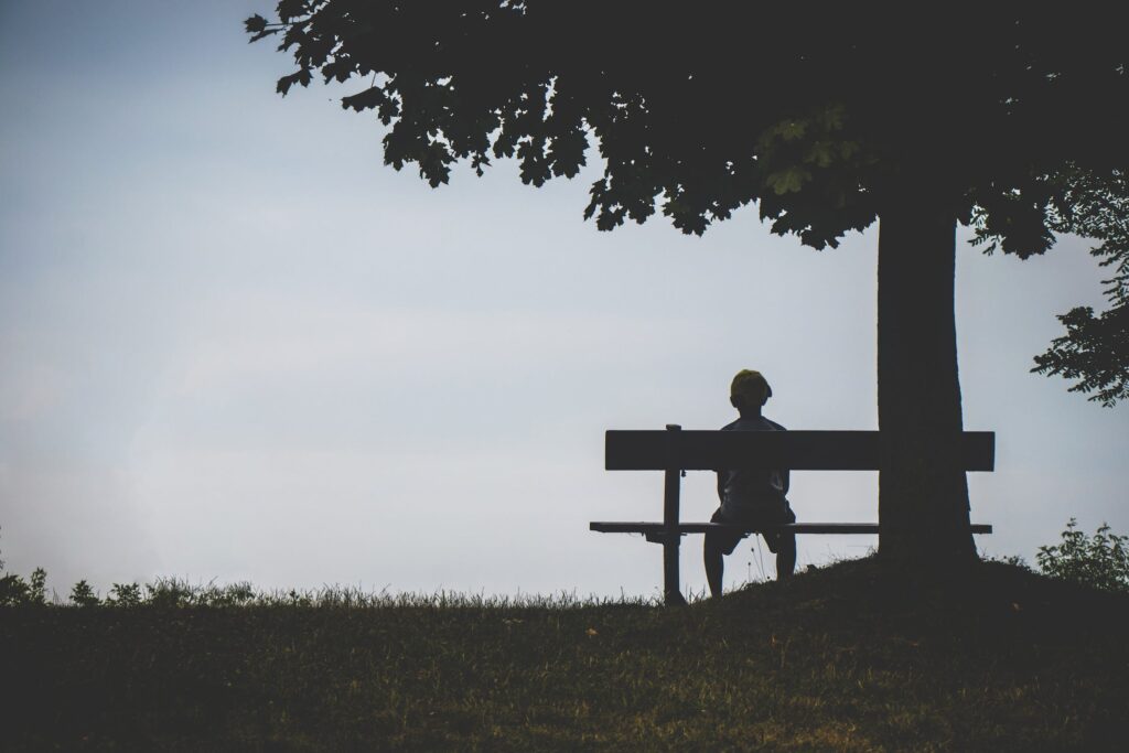 Ein Junge, wecher alleine auf einer Bank unter einem Baum sitzt und in die Ferne schaut.